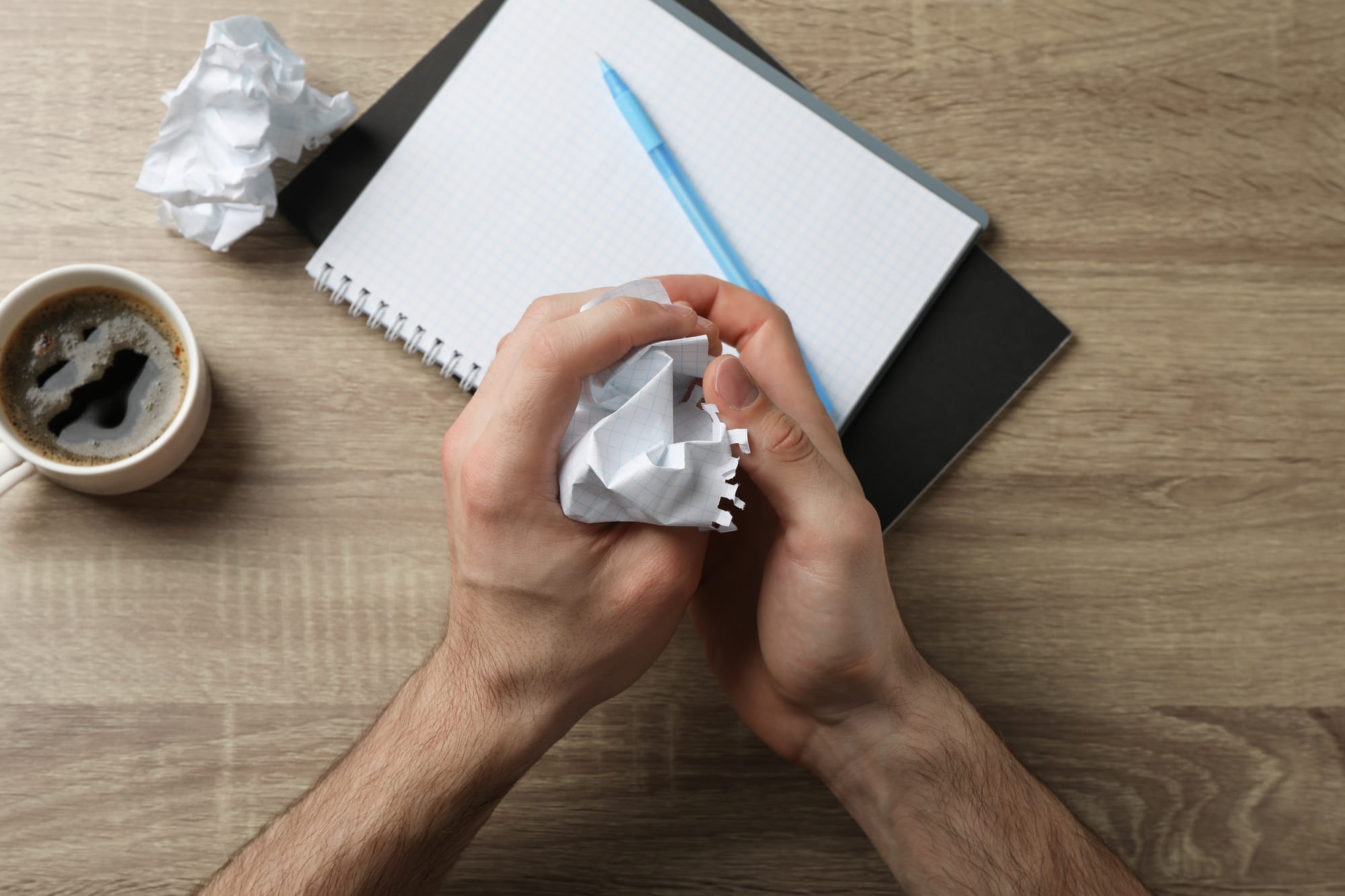 Man crumpling paper on wood table with copybook and cup of coffee, top view